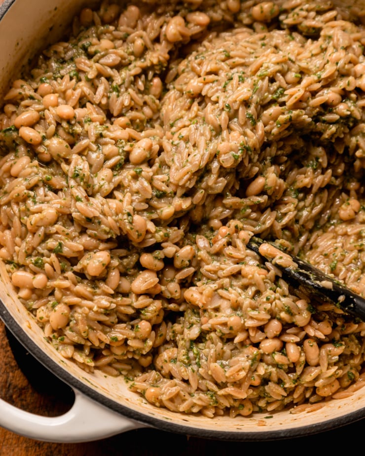 An overhead shot shows a pot filled with cooked orzo pasta, white beans and pesto. The texture is creamy and rich looking.