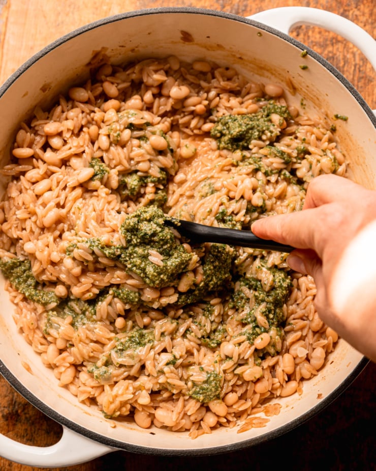 An overhead shot shows a hand using a spatula to stir pesto into cooked orzo pasta and white beans in a pot.