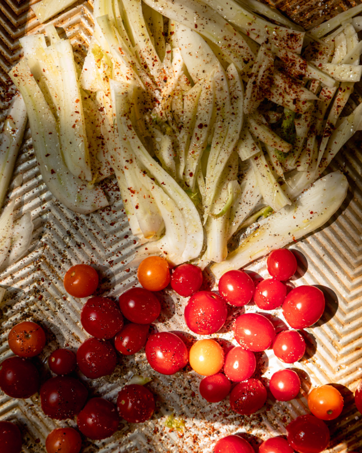 An overhead shot shows sliced fresh fennel and cherry tomatoes on a baking sheet. They have been drizzled with olive oil and seasoned with chili flakes, salt, and pepper.