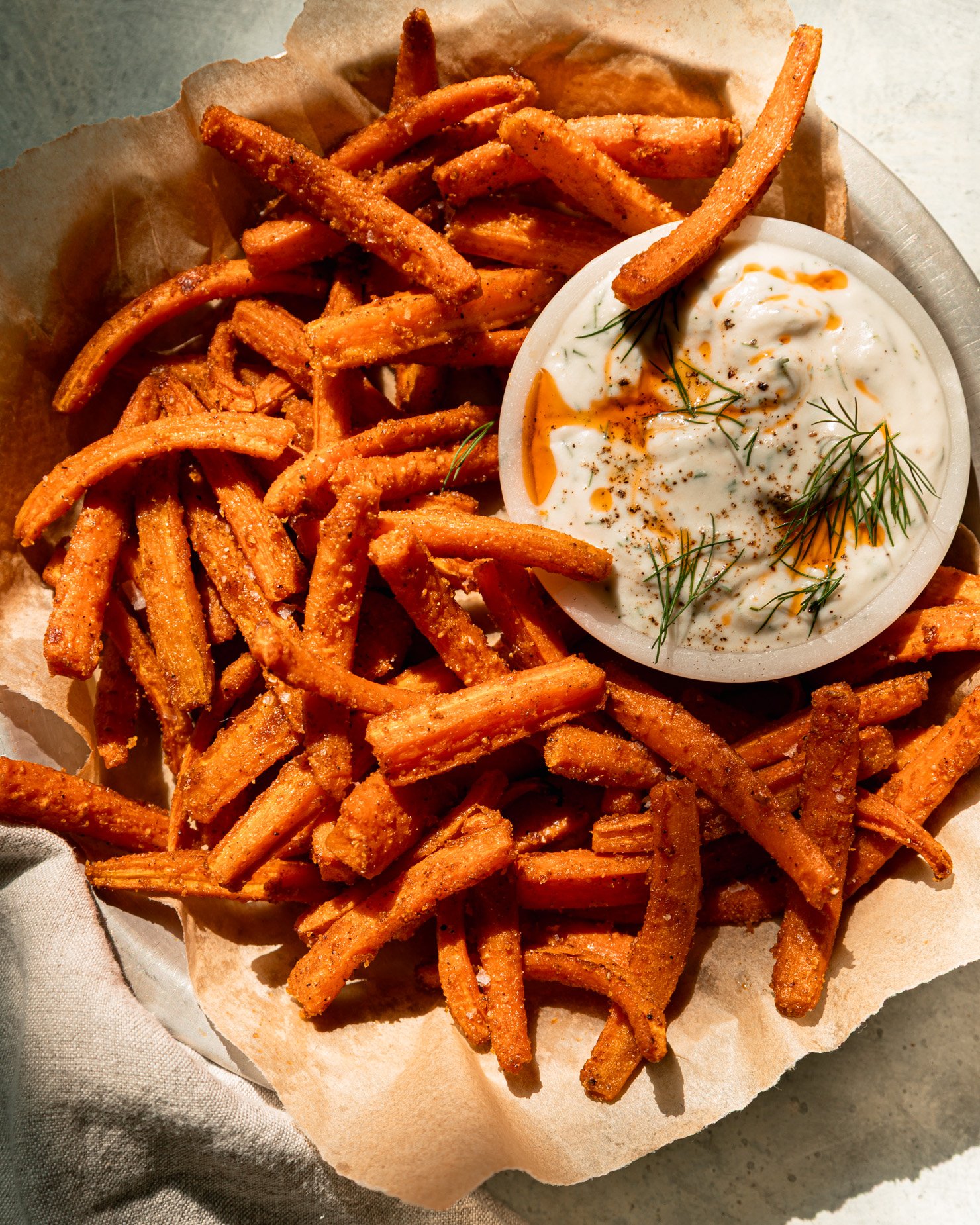 An overhead shot shows a paper-lined dish filled with roasted carrot fries. A small bowl of garlicky dill yogurt dip is seen to the side. The dip is garnished with chili oil and fresh dill. The photo is taken in direct sunlight.
