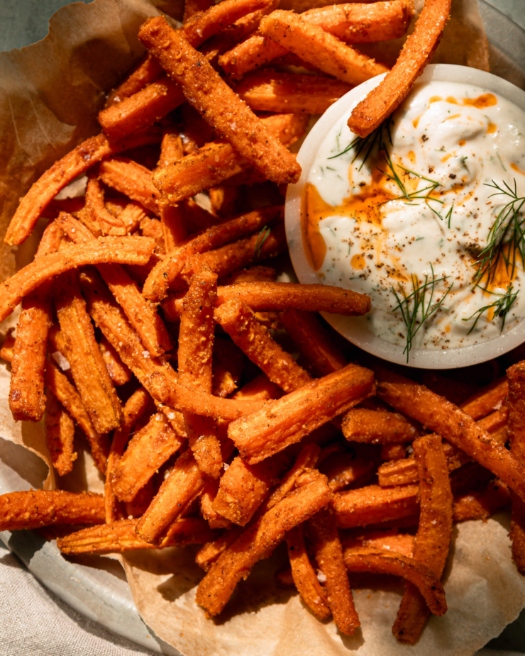 An up close, overhead shot shows a paper-lined dish filled with roasted carrot fries. A small bowl of garlicky dill yogurt dip is seen to the side. The dip is garnished with chili oil and fresh dill. The photo is taken in direct sunlight.