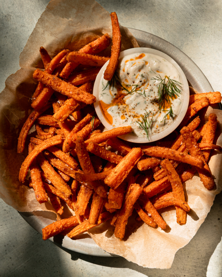 An overhead shot shows a paper-lined dish filled with roasted carrot fries. A small bowl of garlicky dill yogurt dip is seen to the side. The dip is garnished with chili oil and fresh dill. The photo is taken in direct sunlight.