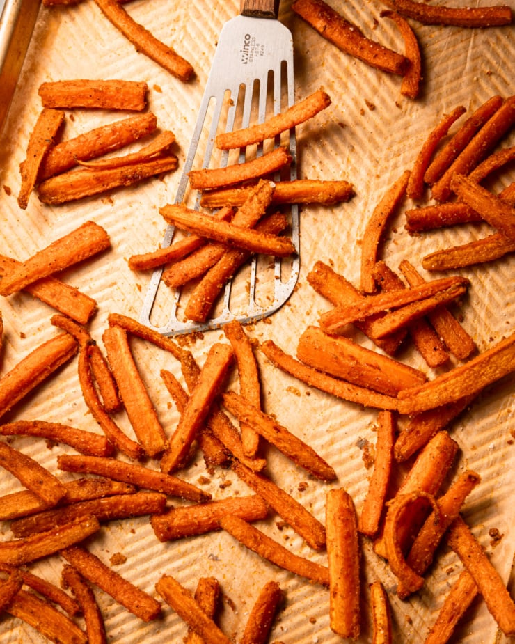 An overhead shot shows roasted carrot fries on a parchment-lined baking sheet. A fish spatula is lifting some off the baking sheet.