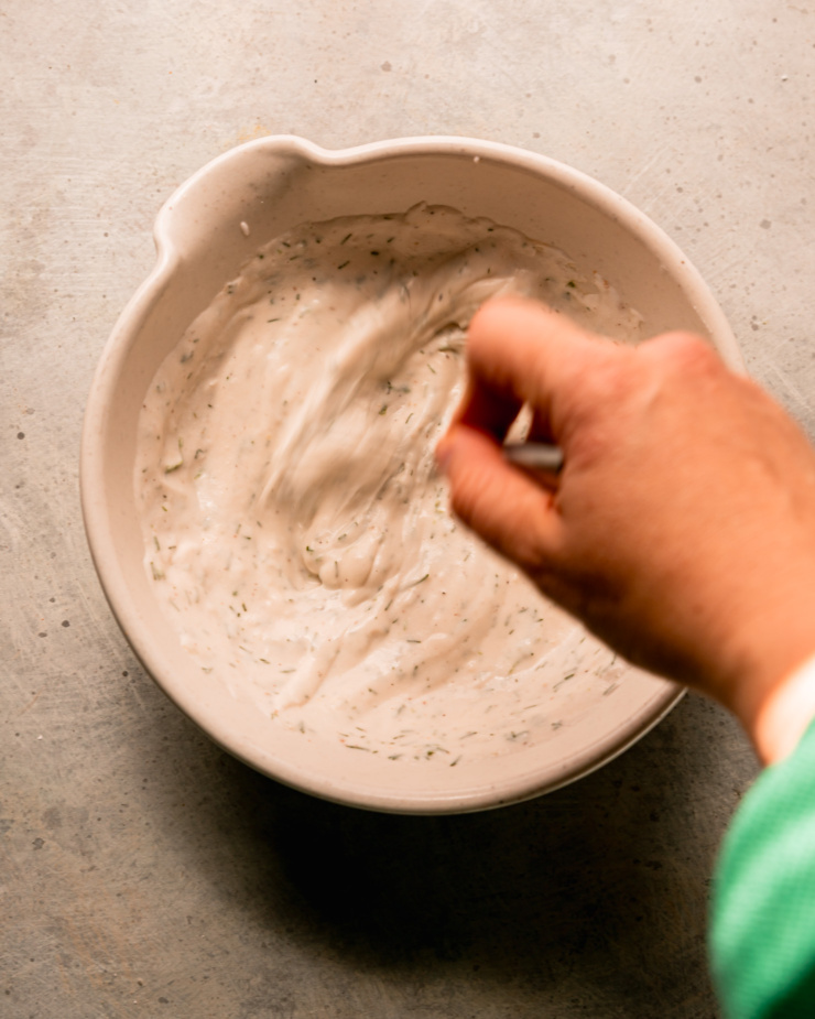 An overhead shot shows a hand whisking a yogurt-based sauce together in a bowl. The whisking motions are a blur.