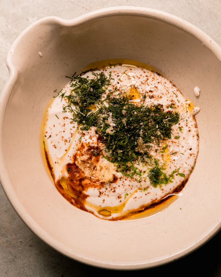 An overhead shot shows a bowl with coconut milk-based yogurt, fresh chopped dill, Tamari, olive oil, lime zest, salt, pepper, and finely minced garlic.