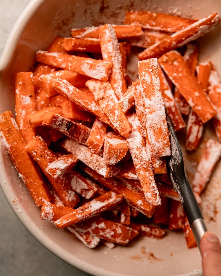 An up close, overhead shot shows a hand using a spatula to stir up cut carrots in a bowl. The carrots are coated in oil, cornstarch, and spices.