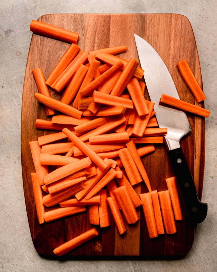 An overhead shot shows a bunch of carrots cut into sticks on a wooden cutting board. A chef knife is seen to the side.