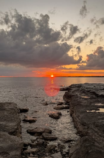 A head-on shot shows a sunset over a lake with a rocky shore in the foreground.