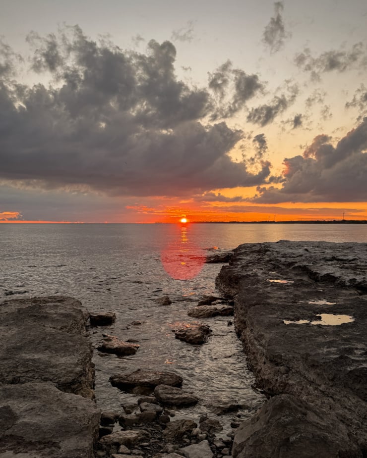 A head-on shot shows a sunset over a lake with a rocky shore in the foreground.