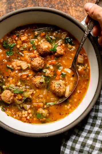 An overhead shot shows an individual bowl of vegan Italian wedding soup with little vegan sausage meatballs, tiny pasta pearls, wilted chopped escarole, and sautéed veggies. The top of the soup is garnished with chopped parsley. A spoon is held by a hand and a checked napkin is nearby.