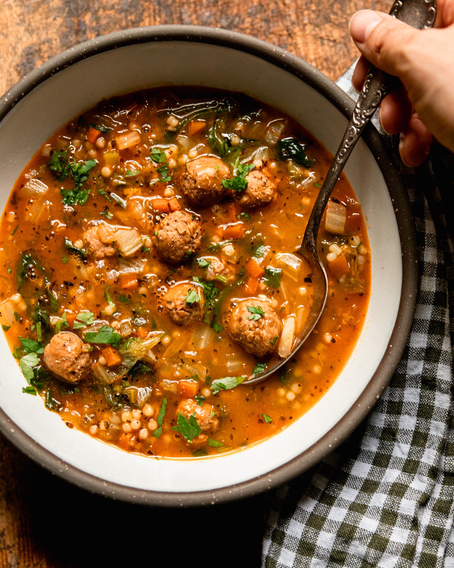 An overhead shot shows an individual bowl of vegan Italian wedding soup with little vegan sausage meatballs, tiny pasta pearls, wilted chopped escarole, and sautéed veggies. The top of the soup is garnished with chopped parsley. A spoon is held by a hand and a checked napkin is nearby.
