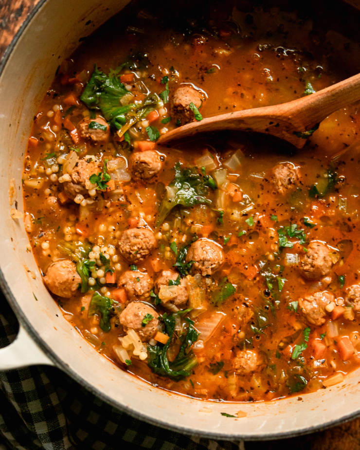 An overhead shot shows a pot of vegan Italian wedding soup. You see wilted escarole, tiny pasta, and little vegan sausage meatballs floating on the surface. The soup is garnished with chopped parsley and a wooden spoon can be seen dipping into the broth.