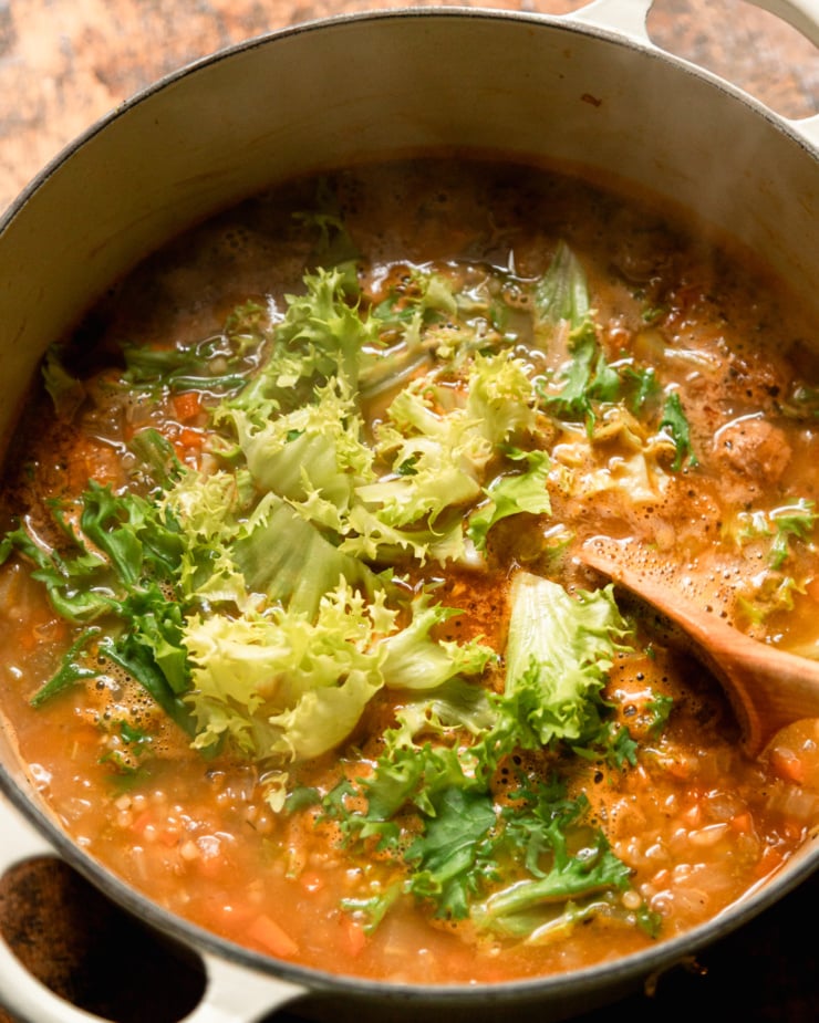 An overhead shot shows chopped escarole that's just been added to a pot of soup. A wooden spoon is in the shot as well.