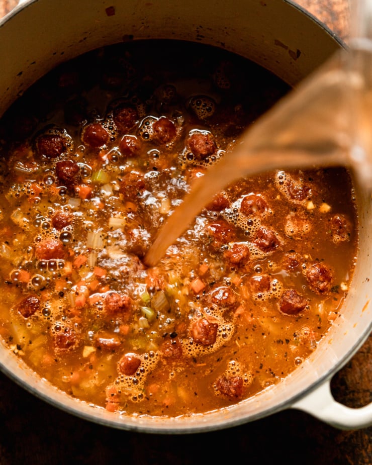 An overhead shot shows vegetable stock being poured into a pot with veggie sausage meatballs and sautéed vegetables.