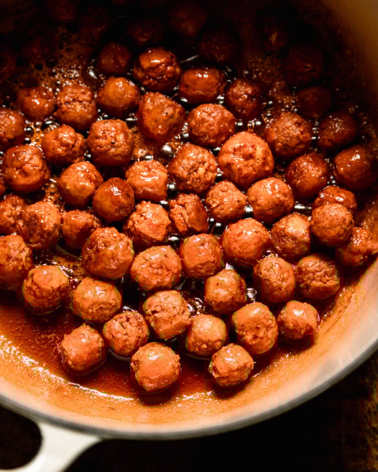 An up close, overhead shot shows sizzling little vegan sausage meatballs in a Dutch oven. They are glistening.