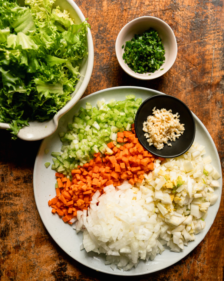 An overhead shot shows a bowl of chopped escarole, a small bowl of chopped parsley, a small bowl of minced garlic, and a dinner plate with chopped fennel, onion, carrots and celery.