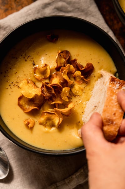 An overhead shot shows a hand dipping a crust of bread into a bowl of creamy vegan parsnip soup. The soup is garnished with parsnip chips.