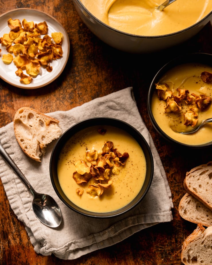 An overhead shot shows 2 individual bowls of parsnip soup garnished with parsnip chips. The plate of parsnip chips and the pot of soup are nearby. Slices of bread are also seen.
