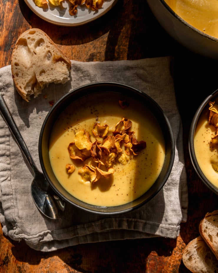 An overhead shot shows an individual serving of parsnip soup garnished with parsnip chips. Bread is nearby and the photo is taken in direct sunlight.