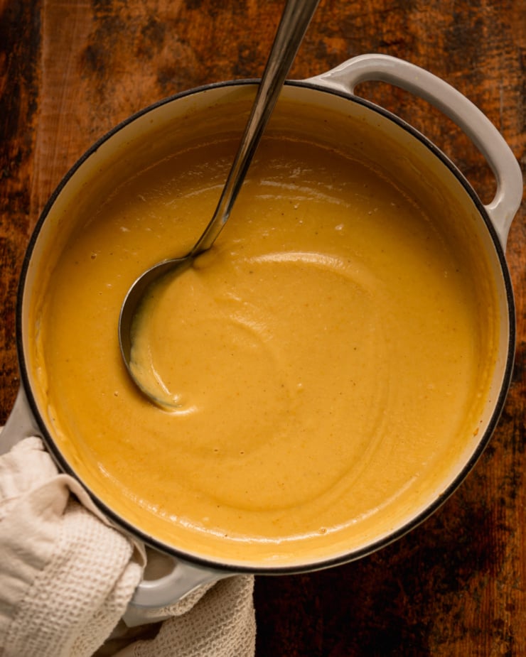 An overhead shot shows a Dutch oven filled with creamy puréed parsnip soup. A ladle is sticking out of the pot.