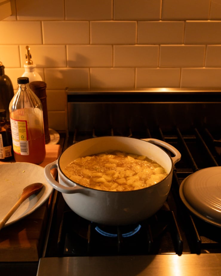 A head-on shot shows a Dutch oven filled with stock and vegetables simmering on a gas stove. A couple bottles and a pepper mill are seen on the nearby counter.