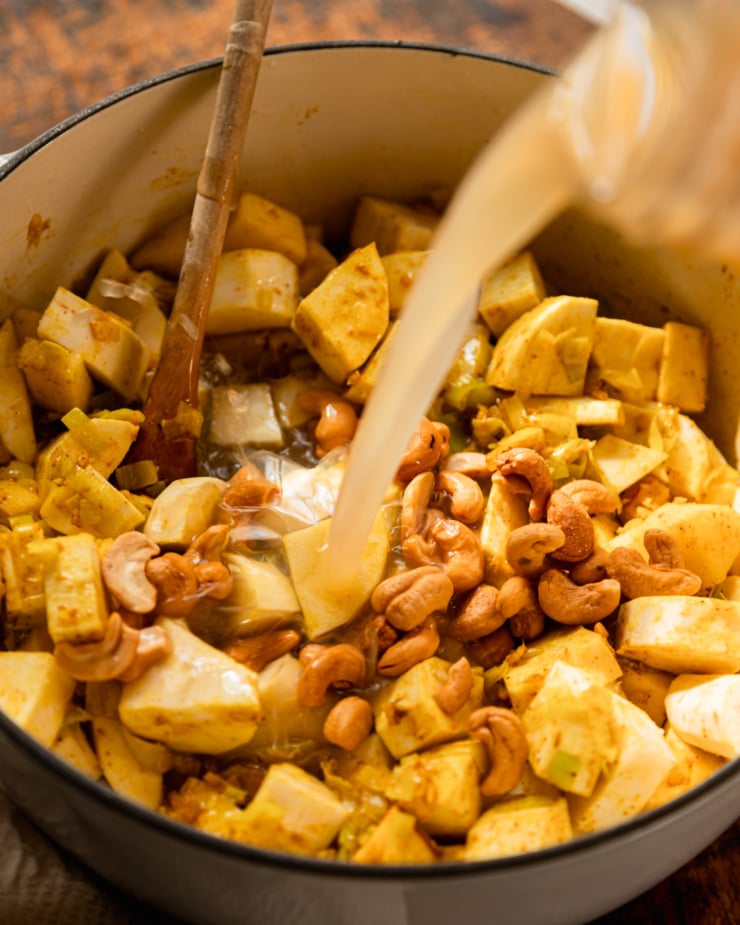 A 3/4 angle shot shows vegetable stock being poured over cooked vegetables and roasted cashews in a pot.