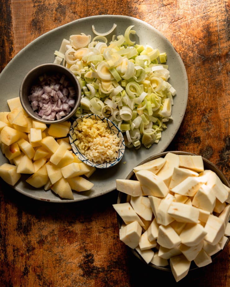 An overhead shot shows sliced leeks, chopped shallots, chopped apples, minced ginger, minced ginger, and chopped parsnips; all on their own plates/in their own bowls as part of prep for a soup.