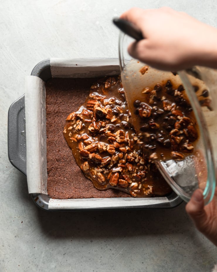 An overhead shot shows a pair of hands pouring a gooey pecan chocolate filling on top of a pan with a baked chocolate crust.