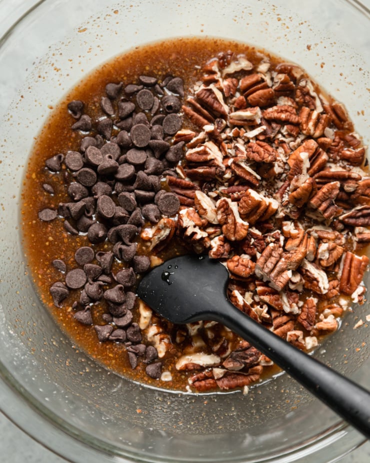 An overhead shot shows a gooey pecan bar filling in a bowl with chopped pecans and chocolate chips on top, about to be folded in.