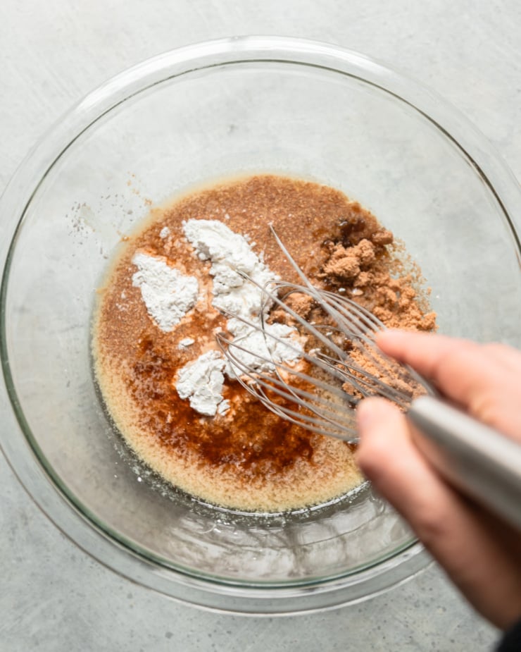 An overhead shot shows a hand holding a whisk in a bowl. In the bowl is brown sugar, melted vegan butter, a flax "egg" mixture, arrowroot starch, maple syrup, vanilla, vinegar, and salt.