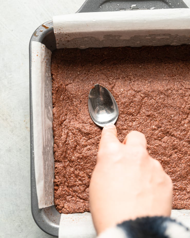An overhead shot shows a baked chocolate crust in a square pan. A hand is using the back of a spoon to press the crust down and firm it up a bit.