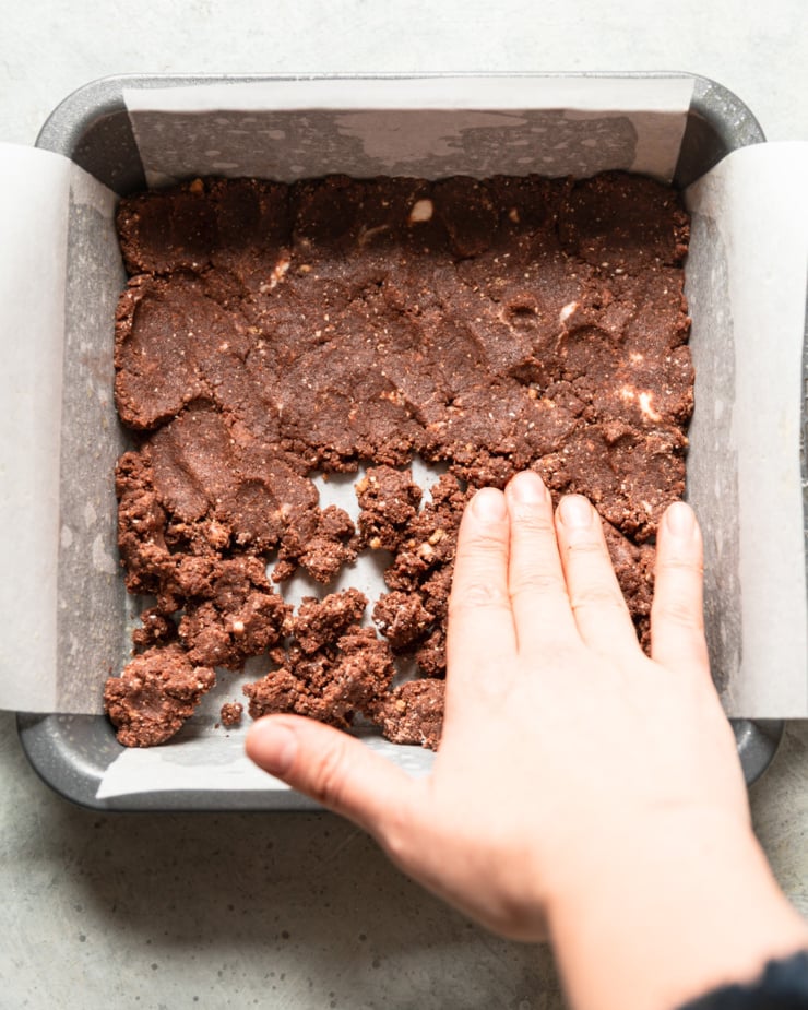 An overhead shot shows a hand pressing a chocolate crust mixture into a parchment lined pan.