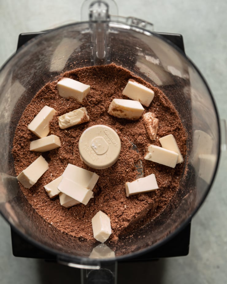 An overhead shot shows a cocoa and dry flour mixture in a food processor with bits of cold vegan butter on top.