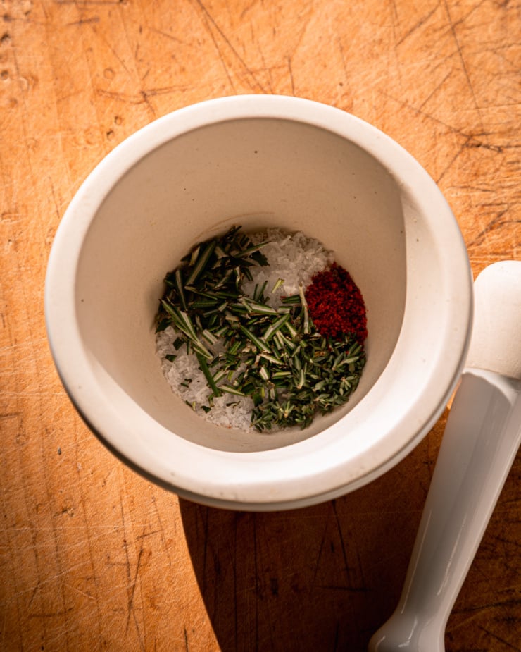 An overhead shot shows a mortar and pestle with some flaky salt, herbs, and ground chili in the mortar.