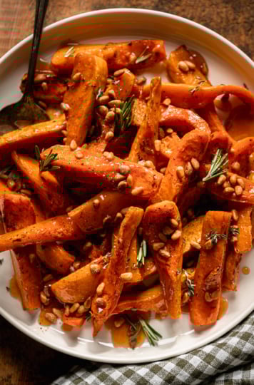 An overhead shot shows roasted honeynut squash wedges on a plate, topped with toasted pine nuts, herbs, and apple cider reduction. A check linen napkin is nearby and a silver serving spatula is sticking out of the squash.