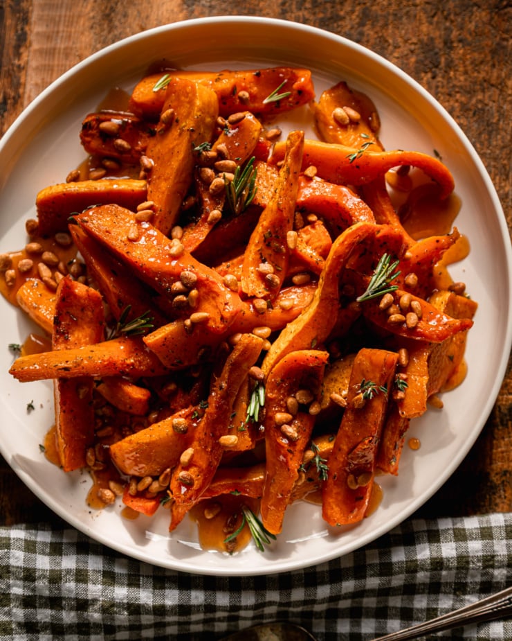 An overhead shot shows roasted honeynut squash wedges on a plate, topped with toasted pine nuts, herbs, and apple cider reduction. A check linen napkin is nearby.