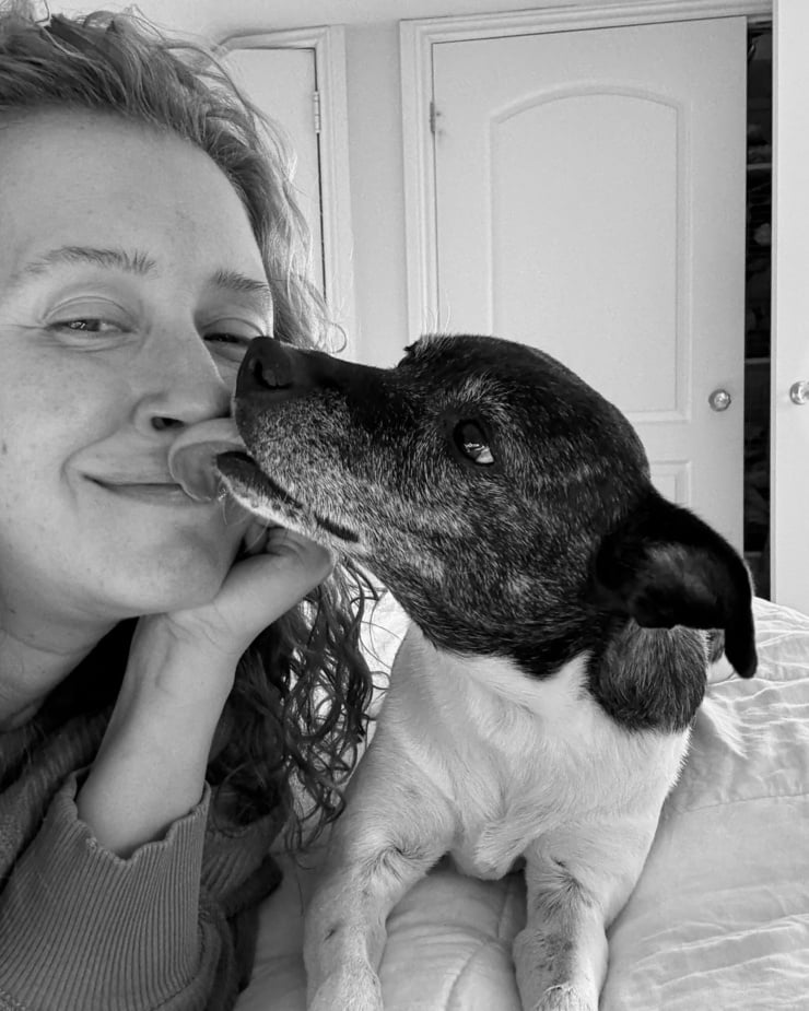A selfie shot shows a woman and her jack russell hound mix dog. The dog is licking her face.