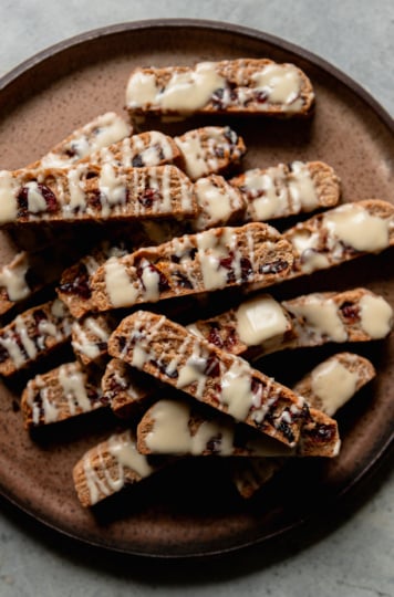 An overhead shot shows a plate piled up with vegan orange cranberry biscotti, all drizzled with white chocolate.