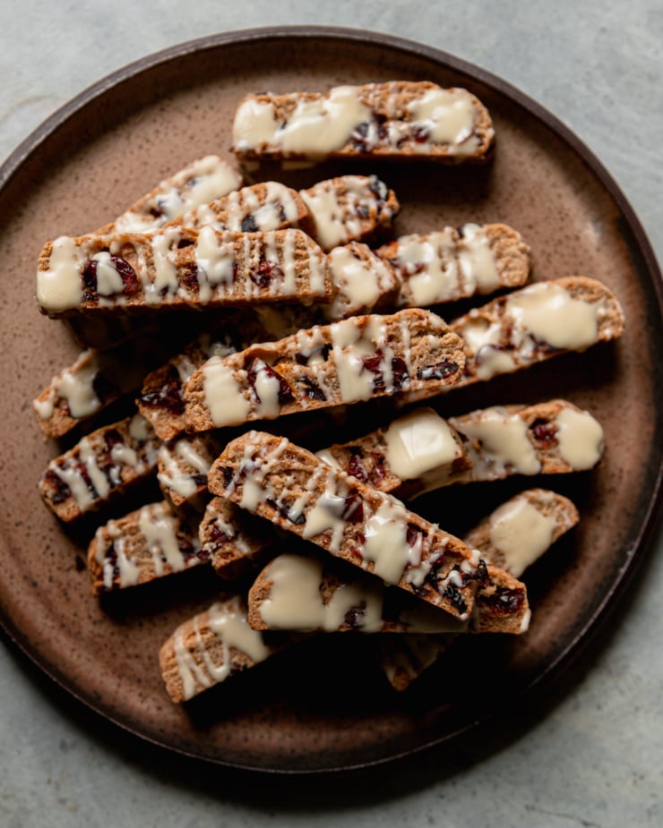 An overhead shot shows a plate piled up with vegan orange cranberry biscotti, all drizzled with white chocolate.