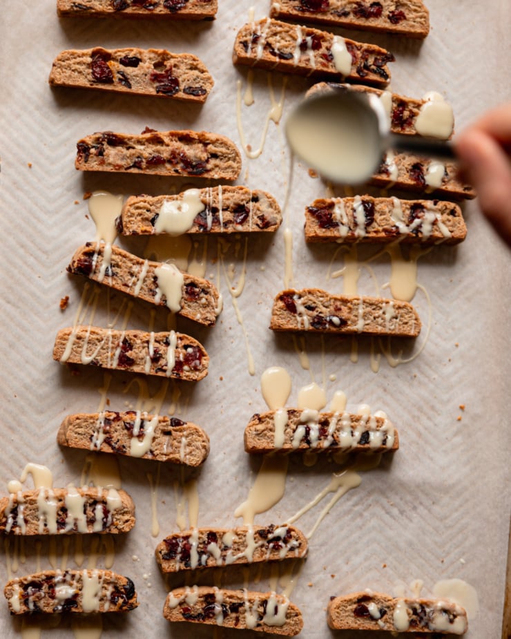 An overhead shot shows a hand drizzling white chocolate with a spoon over vegan orange cranberry biscotti.