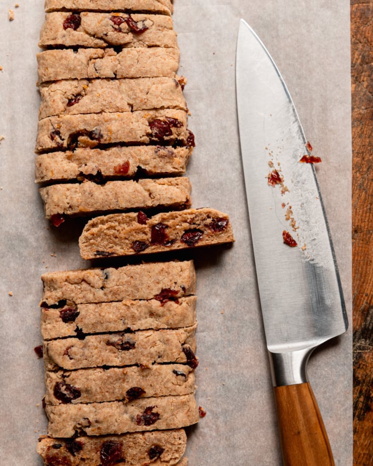 An overhead shot shows biscotti being sliced after their first bake. A chef's knife is seen to the side.