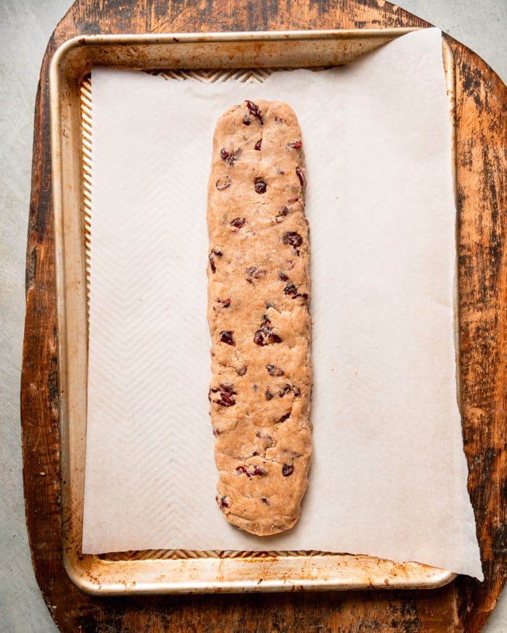 An overhead shot shows biscotti dough shaped into a log on a baking sheet, ready for its first bake.