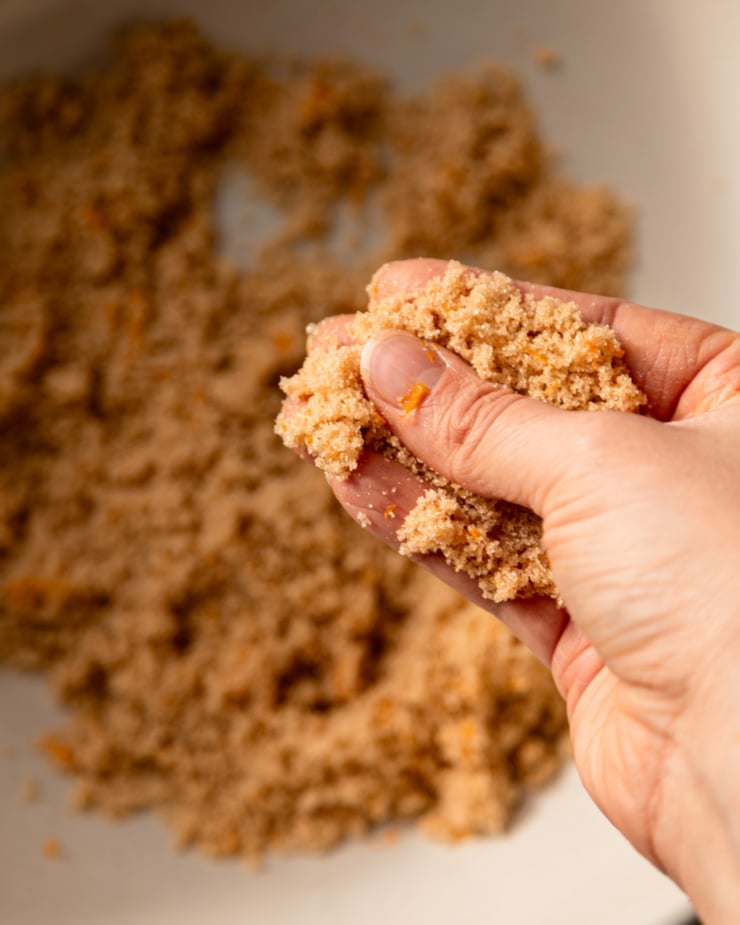 An overhead shot shows a hand working orange zest into some light brown sugar.