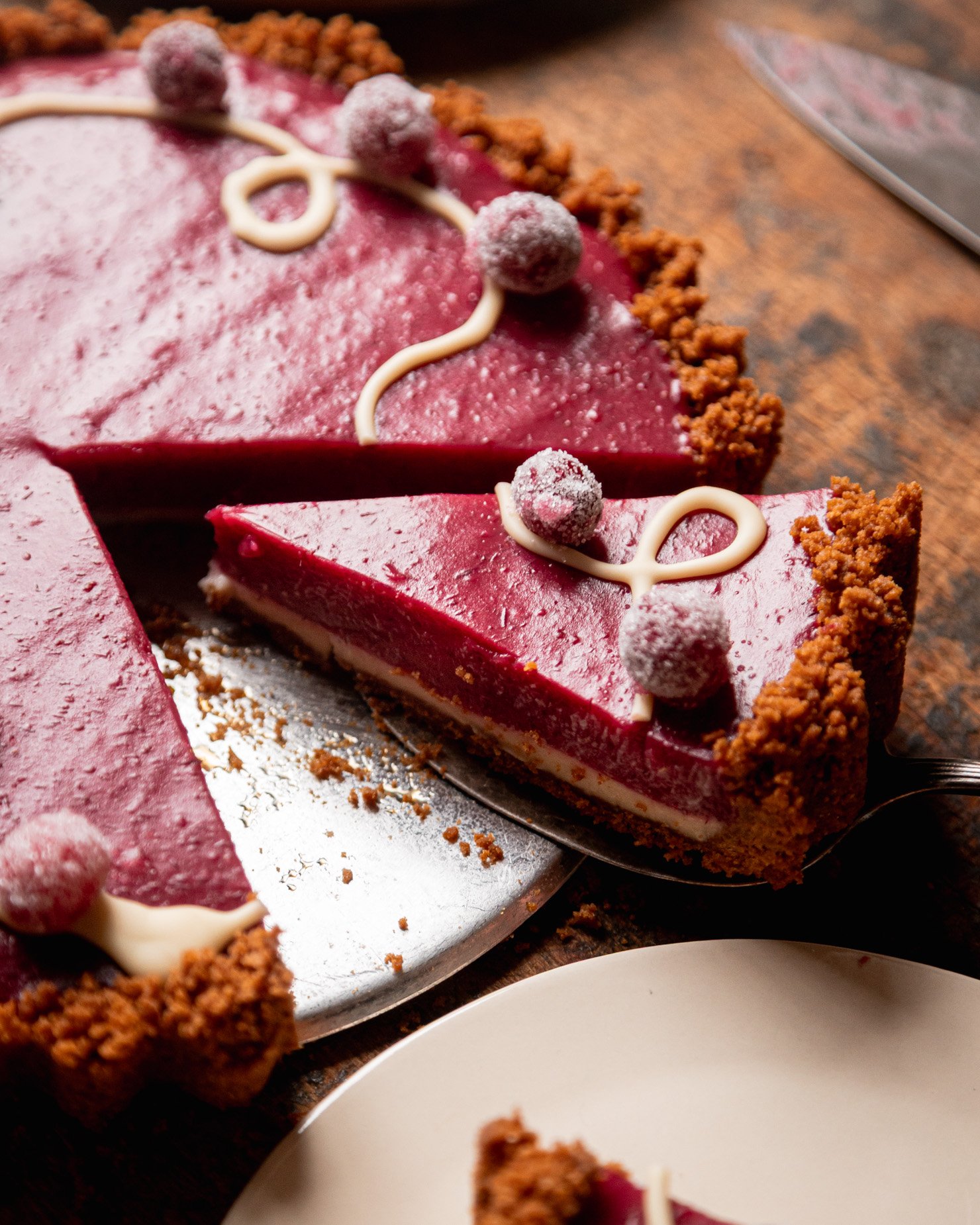A 3/4 angle image shows a slice being taken out of a vegan cranberry tart. You can see a defined white chocolate bottom layer with the cranberry curd layer on top. The top is garnished with melted white chocolate and sugared cranberries.