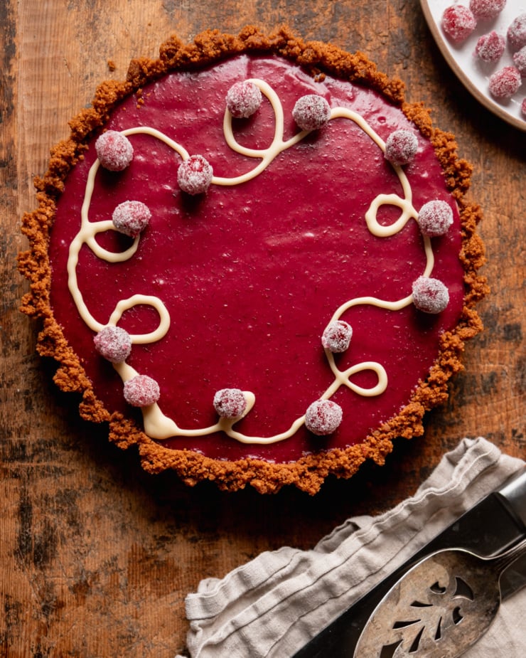 An overhead shot shows a vegan cranberry tart with a white chocolat5e drizzle and sugared cranberries on top. A knife, pie server, and napkin are nearby.