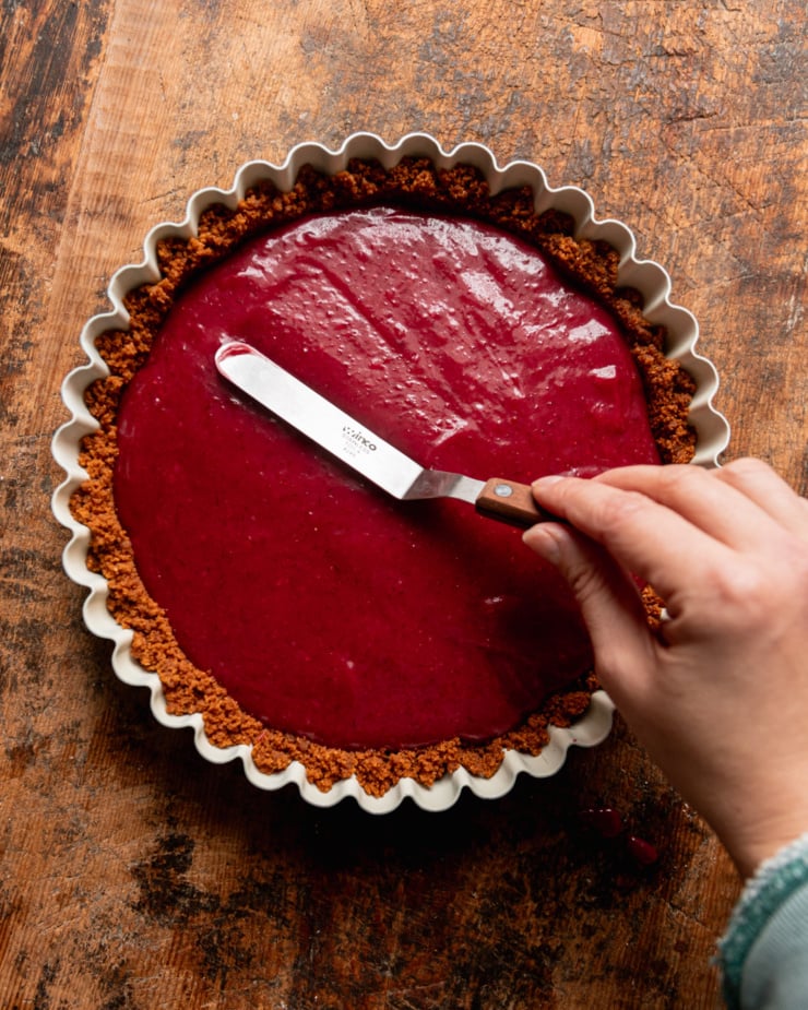 An overhead shot shows a hand using a small offset spatula to smooth out a vegan cranberry curd filling over a tart.