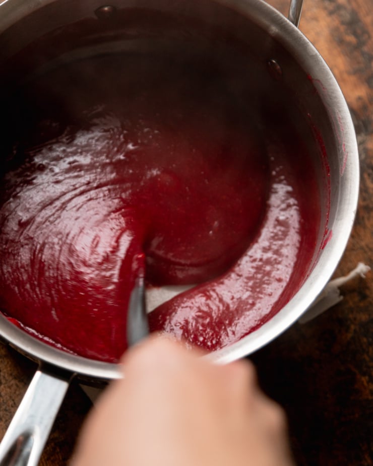 An overhead shot shows a hand using a spatula to stir up some vegan cranberry curd in a saucepan.