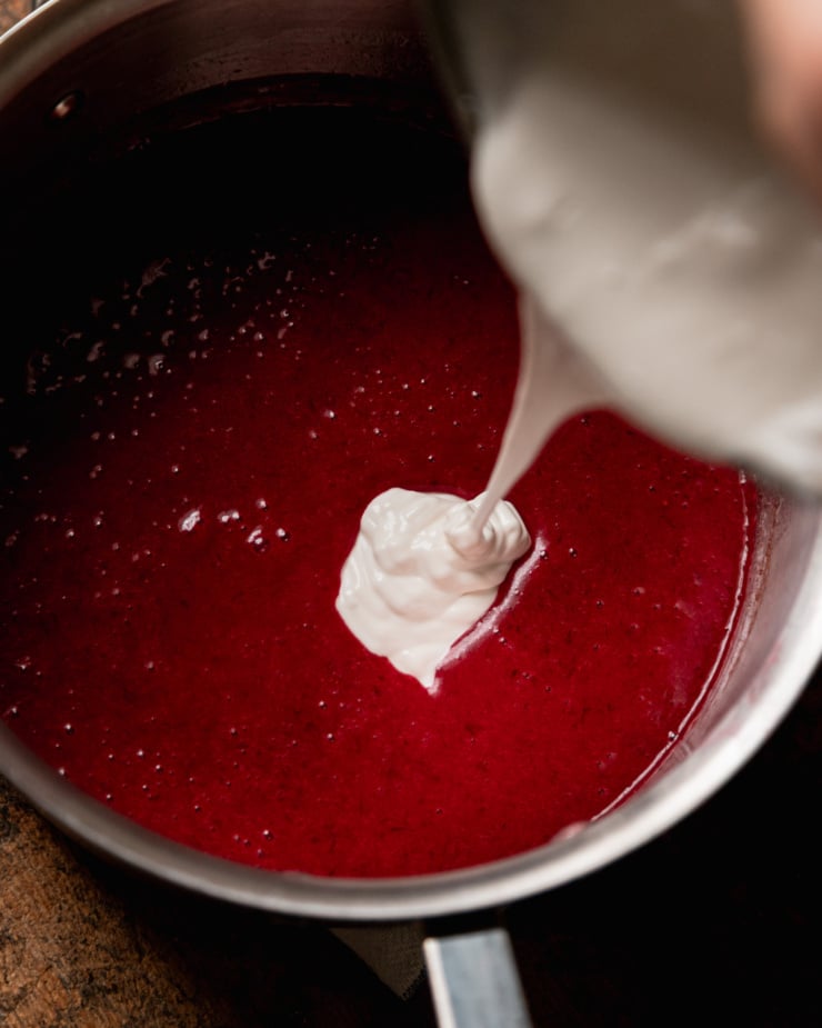 An overhead shot shows a coconut yogurt and cornstarch slurry being poured into a puréed cranberry mixture in a saucepan.