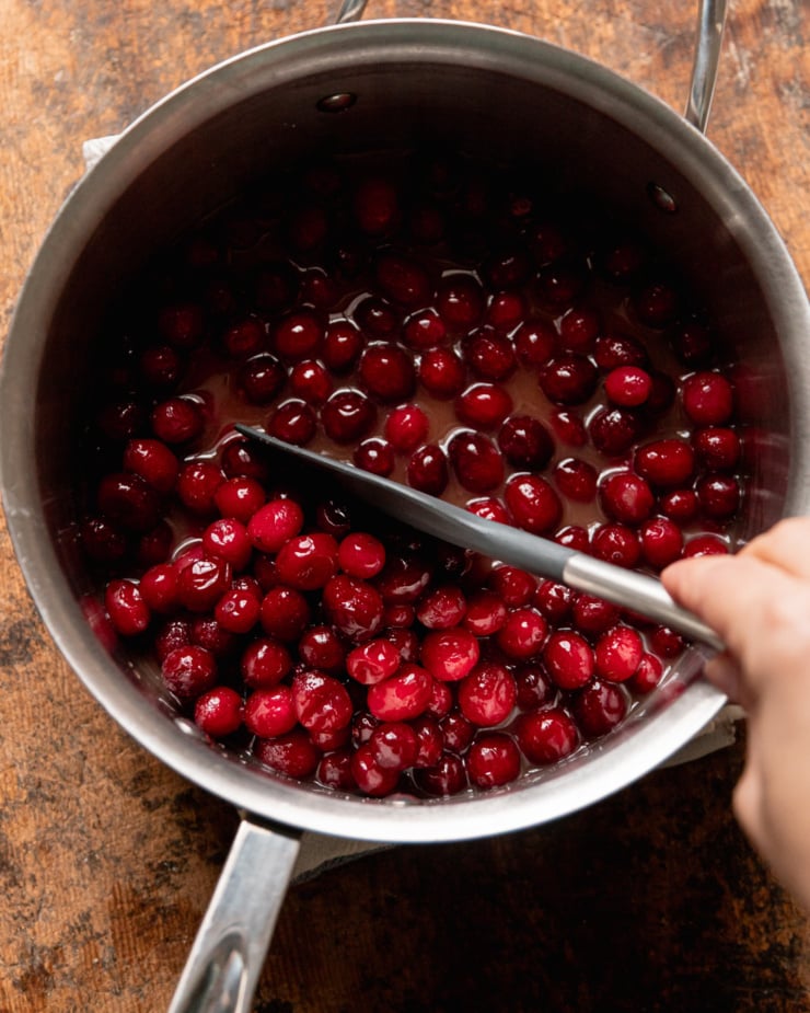 An overhead shot shows whole cranberries in a liquid mixture in a saucepan. A hand is stirring them with a spatula.
