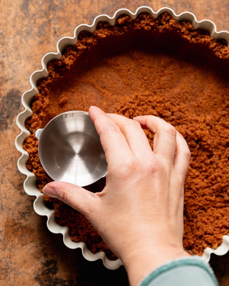 An overhead shot shows a hand using a dry measuring cup to firm in a Biscoff cookie crust mixture into a tart pan.